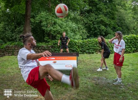 fotografia. dni wyszehradzkie w willi decjusza. mecz teqball na wolnym powietrzu. czworo zawodników wokół stołu ustawionego na trawie. w tle budynek willi decjusza. mężczyzna podnosi wysoko nogę do kopięcia.